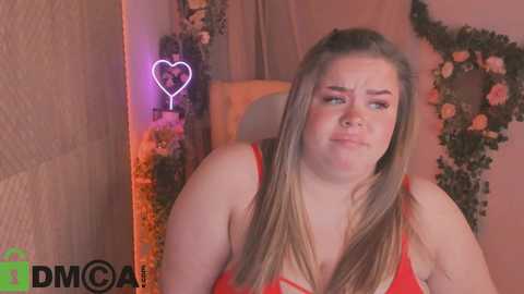 Media: Video of a plus-sized, fair-skinned woman with long blonde hair in a red tank top, sitting in a cozy room with floral decorations and a glowing heart-shaped neon sign.