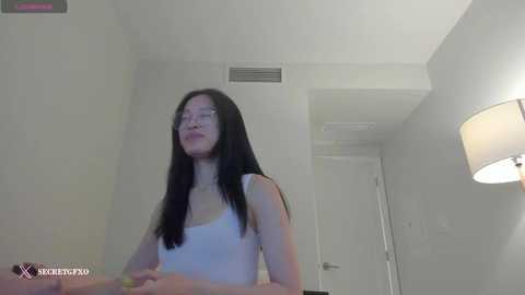 Media: A video of a young Asian woman with long black hair, wearing glasses and a white tank top, smiling and looking upwards, in a minimalist, white-walled room with a closed door and a lamp.