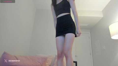 Media: Video of a slender, light-skinned woman with long dark hair, wearing a black crop top and mini skirt, standing in a bright, minimalist room with white walls and a pink couch.