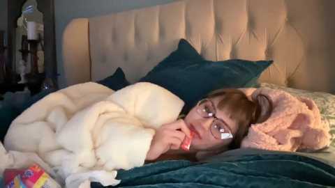 Media: A video of a young woman with glasses and light brown hair, wrapped in a white blanket, lying on a bed with teal sheets and pillows.