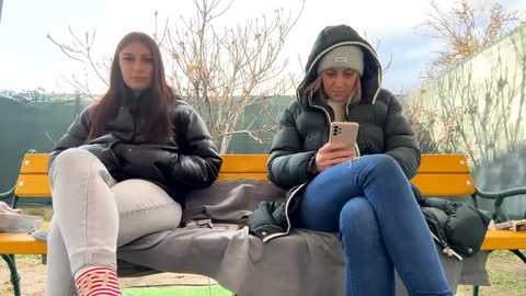 Media: Video of two young women, one in a black jacket, white pants, and red socks, the other in a dark hoodie and blue jeans, sitting on a yellow bench in a park.