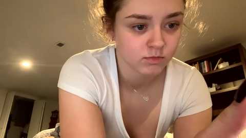 Media: Video of a young, fair-skinned woman with light brown hair tied back, wearing a white top, looking thoughtful, in a dimly lit room with a bookshelf and a lamp in the background.