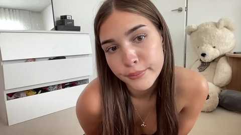 Media: Video of a young woman with long brown hair, fair skin, and natural makeup, wearing a necklace, kneeling in a minimalist room with white furniture and a plush teddy bear.