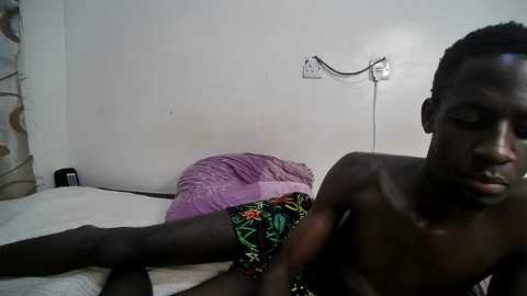 Media: Video of a dark-skinned, shirtless young man with a lean physique lying on a bed with a purple pillow and floral-patterned shorts, in a simple, dimly lit room.