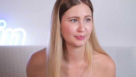 Media: Video of a fair-skinned, blonde-haired woman with light blue eyes, wearing minimal makeup, smiling slightly, with a white textured background and blurred neon signs.