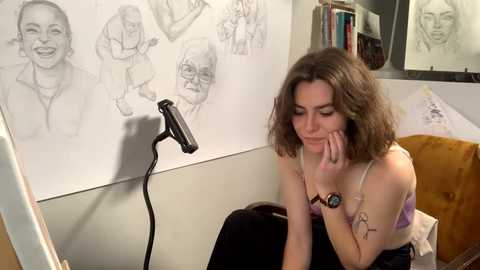 Media: Video of a young woman with curly brown hair, wearing a purple bra, seated at a desk with a drawing tablet, surrounded by sketchbook pages and a lamp.