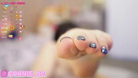 Media: A close-up video of a woman's bare foot, toenails painted in metallic silver, with a blurred background featuring a pink and white patterned bedspread.