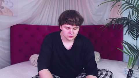 Media: Video of a young Caucasian man with short brown hair, wearing a black T-shirt, lying on a bed with a pink headboard and white sheets. Background includes a potted plant and draped fabric.