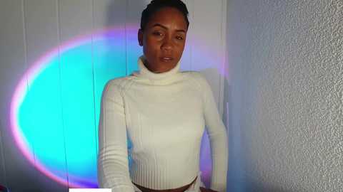 Media: Video of a woman with medium brown skin, dark hair in a bun, wearing a white ribbed turtleneck, standing against textured white wall with a blue and purple circular light behind her.