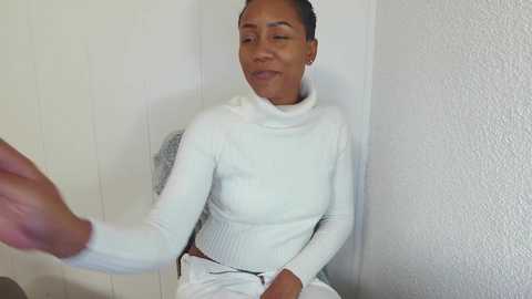 Media: A video of a smiling Black woman in a white turtleneck and pants, sitting against a textured white wall, with a hand extending into the frame.