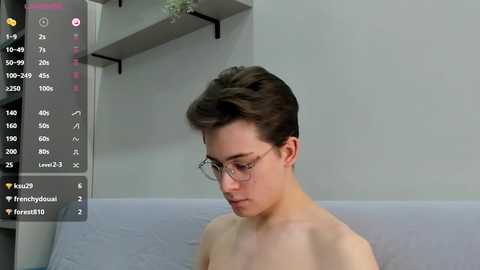 Media: Video of a fair-skinned, short-haired, young, shirtless man with glasses, sitting on a light gray couch. Background includes a white wall with a shelf and potted plant.