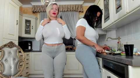 Media: Video of two women in a modern kitchen: one blonde, one brunette, both in tight, white tops and light grey leggings, preparing food.