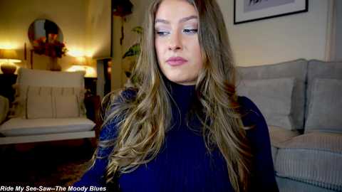 Media: Video of a young woman with long, wavy blonde hair, wearing a dark blue sweater, standing in a cozy, warmly lit living room with beige furniture and a framed picture on the wall.