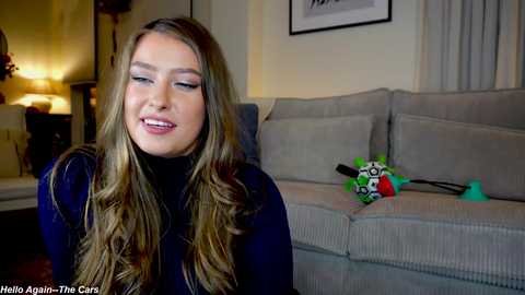 Media: Video of a smiling, light-skinned woman with long, wavy blonde hair, wearing a navy blue turtleneck, sitting on a beige sofa in a cozy living room. A colorful plush toy is nearby.