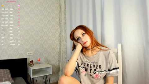 Media: Video of a pale-skinned, red-haired woman in a gray \"Forever NYC\" T-shirt, leaning on a bed, with a white wall calendar behind her.