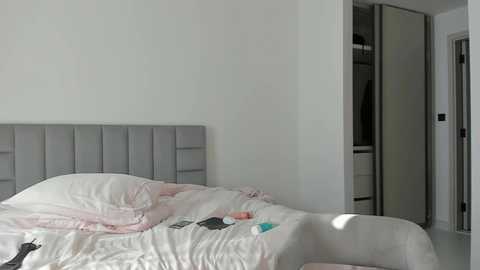 Media: Video of a minimalist bedroom with a gray tufted headboard, white bedding, and a partially open wardrobe revealing a hanging coat.