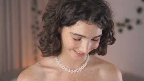 Media: Video of a woman with curly dark hair, wearing a pearl necklace, looking down with a gentle smile. Background features blurred floral wallpaper.