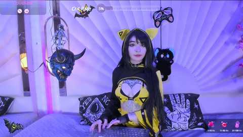 Media: Video of a woman in a black and yellow costume, resembling a witch, seated at a desk in a dimly lit room with purple lighting and various Halloween decorations.