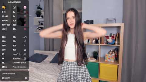 Media: Video of a young woman with long dark hair, wearing a white crop top and plaid skirt, standing in a modern bedroom with a wooden shelf, colorful storage bins, and a bed.