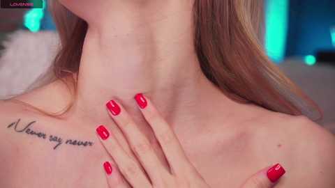 Media: A close-up video of a fair-skinned woman with long, straight brown hair. She has a tattoo on her left shoulder and her right hand, with red-painted nails, gently touches her neck. The background is blurred, featuring blue and white lighting.