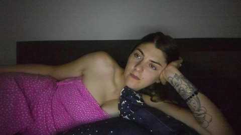 Media: Video of a woman with fair skin, dark hair, and tattoos on her arm, lying on a bed in a dimly lit room, wearing a pink polka-dot dress.