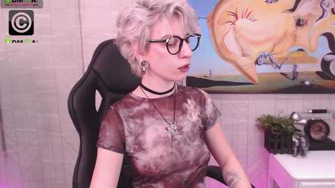 Media: Video of a pale-skinned woman with short, platinum blonde hair, wearing black-framed glasses, a tie-dye top, and black choker. She sits on a black gaming chair, with a colorful mural and potted plants in the background.