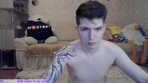 Media: Video of a young, shirtless man with short brown hair and a large tattoo on his left arm, sitting in a cluttered, beige room with stuffed animals and a \"MOSCOW\" shirt.