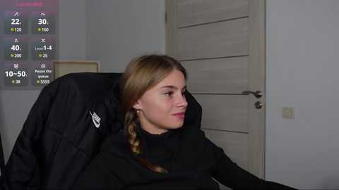 Media: Video of a young Caucasian woman with light skin and blonde hair in a black turtleneck sweater, sitting in a black chair, indoors with a white door and digital clock displaying 10:50 AM.