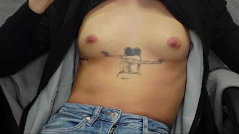 Media: Video of a light-skinned, topless woman with small breasts, wearing a black jacket, and blue jeans, displaying a tattoo of a naked woman with a strap-on dildo between her legs.