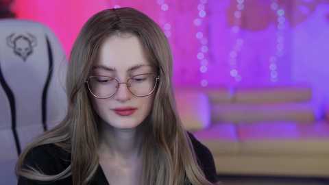 Media: Video of a young woman with long blonde hair, wearing glasses and a black top, seated in a dimly lit room with purple and pink LED lights in the background.