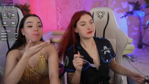 Media: Video of two women, one with long black hair in a gold sequin dress, the other with red hair in a black shirt, sitting on gaming chairs in a dimly lit room.