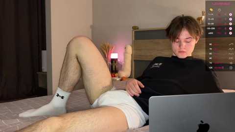 Media: Video of a young, pale-skinned man with a slender build, wearing white Under Armour shorts and a black shirt, lying on a bed with an Apple laptop on his lap.