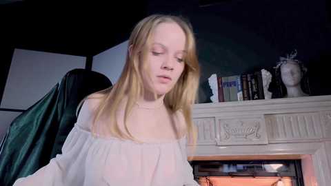 Media: Video of a young Caucasian woman with light blonde hair, wearing a white off-the-shoulder top, standing in front of a white fireplace adorned with books and a bust sculpture.