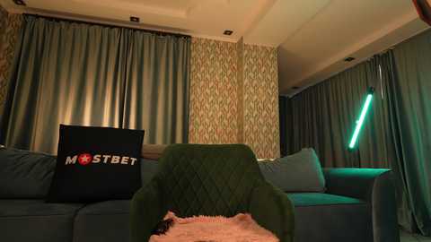 Media: A video of a dimly lit living room with dark green couches, a neon green lamp, patterned wallpaper, and a \"MOSTBET\" pillow.