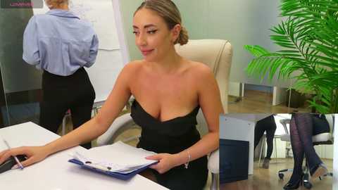 Media: Video of a light-skinned woman with blonde hair in a black strapless dress, seated at a desk, holding a clipboard, in a modern office with green plants, whiteboard, and another person in the background.