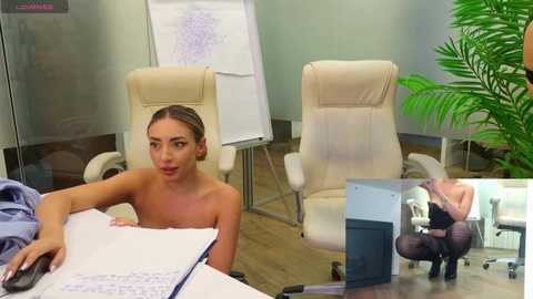 Media: Video of a woman with light skin, brown hair, and medium-sized breasts, topless, sitting at a desk, surrounded by a modern office with white chairs and green plants.