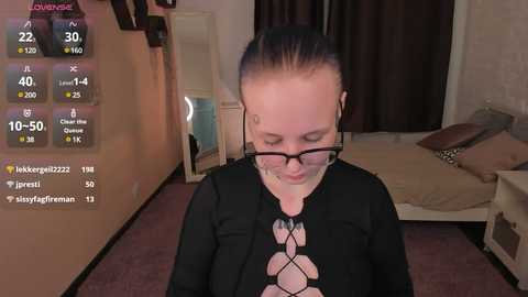 Media: Video of a light-skinned woman with dark hair in a ponytail, wearing glasses and a black, patterned top, standing in a dimly-lit bedroom with a bed and digital health monitoring displays.