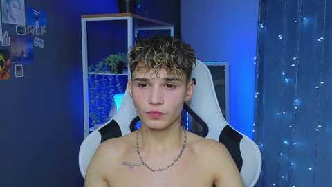 Media: Video of a shirtless young man with curly brown hair, wearing a silver chain necklace, sitting on a white gaming chair in a dimly lit room with blue lighting, posters, and string lights in the background.