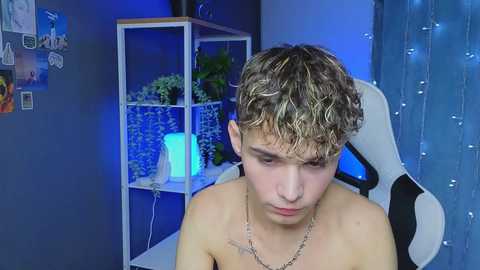 Media: Video of a shirtless, light-skinned, curly-haired teenage boy with a neutral expression in a dimly lit room with blue lighting, plants, and a white gaming chair.