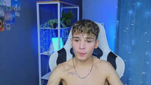 Media: Video of a shirtless, curly-haired young man with light skin and a small tattoo on his chest, seated in a black-and-white gaming chair, surrounded by blue lights, a potted plant, and a lit lamp in a dimly lit room.