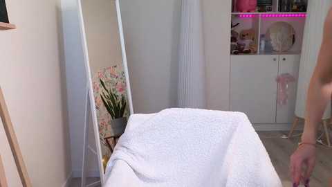 Media: A video of a serene, minimalist spa room with a white massage table covered in a textured blanket. The walls are white, adorned with floral prints and potted plants. A tall mirror reflects the space, and a shelving unit with soft pink lighting is visible.