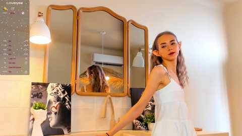 Media: Video of a young woman with long, light brown hair, wearing a white dress, standing near a wooden-framed mirror with a reflection of her. Background features a white wall, a calendar, and a potted plant.