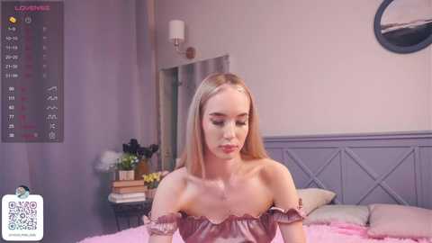 Media: Video of a fair-skinned, blonde woman in a pink off-shoulder dress, sitting on a bed in a softly lit, modern bedroom.