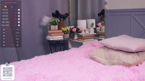 Media: Video of a cozy bedroom with a fluffy pink rug, beige pillows, a stack of books, and floral arrangements on a white nightstand.