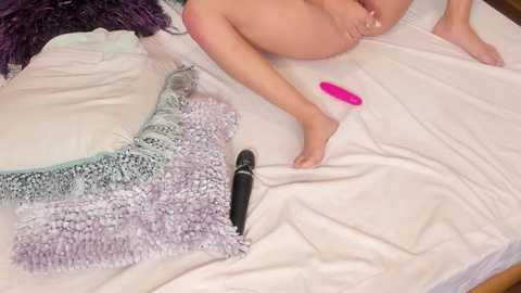 Media: Video of a nude woman sitting on a white bed, legs spread, holding a pink vibrator. Nearby, a black dildo and a lacy purple blanket are visible.