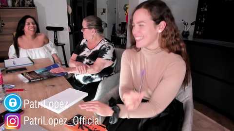 Media: Video of three women laughing around a table; one in a white off-shoulder top, one in a black tie-dye top, and one in a beige top. Background shows a modern living room with a black grand piano.