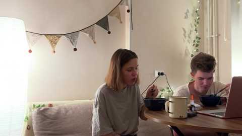 Media: Video of a cozy living room with a woman in a grey top eating popcorn and a man in a striped shirt playing a video game, both seated at a wooden table.