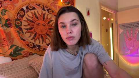 Media: A video of a young woman with straight brown hair, wearing a gray shirt, sitting in a bedroom with a colorful, sun-themed tapestry and a door with a peace sign poster.