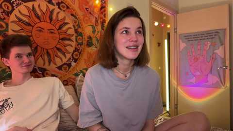 Media: Video of a young couple with a relaxed, cozy vibe. The woman with shoulder-length brown hair, wearing a gray shirt, sits on a couch. Behind them is a sun tapestry and a colorful poster.