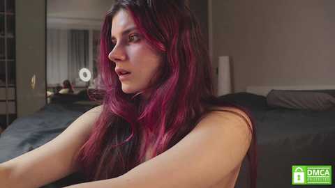 Media: Video of a fair-skinned woman with long, straight, dark purple hair, wearing a sleeveless top, sitting in a modern bedroom with a bed and a mirror.